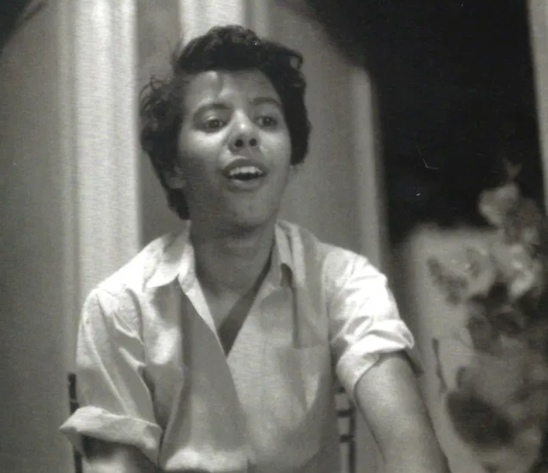 Black and white photograph of Lorraine Hansberry, taken by Molly Cook. Hansberry has her mouth open in the middle of a song. Biographer Imani Perry noted the unique intimacy and personality captured in this candid photo of Hansberry by one of her lovers.
